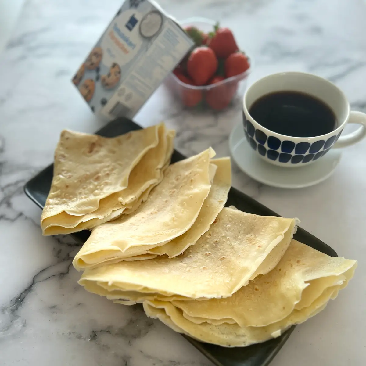 4 crepes folded on a dark blue tray on a table, served with coffee. A powdered sugar package and a box with fresh strawberries are on the table.