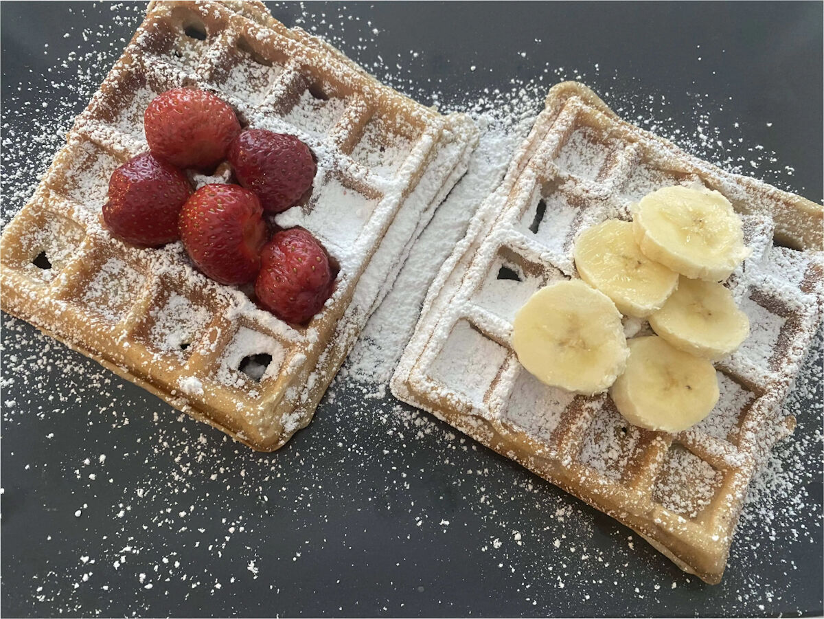 Belgian waffles, one with strawberries and powdered sugar and a second with banana slices and powdered sugar.