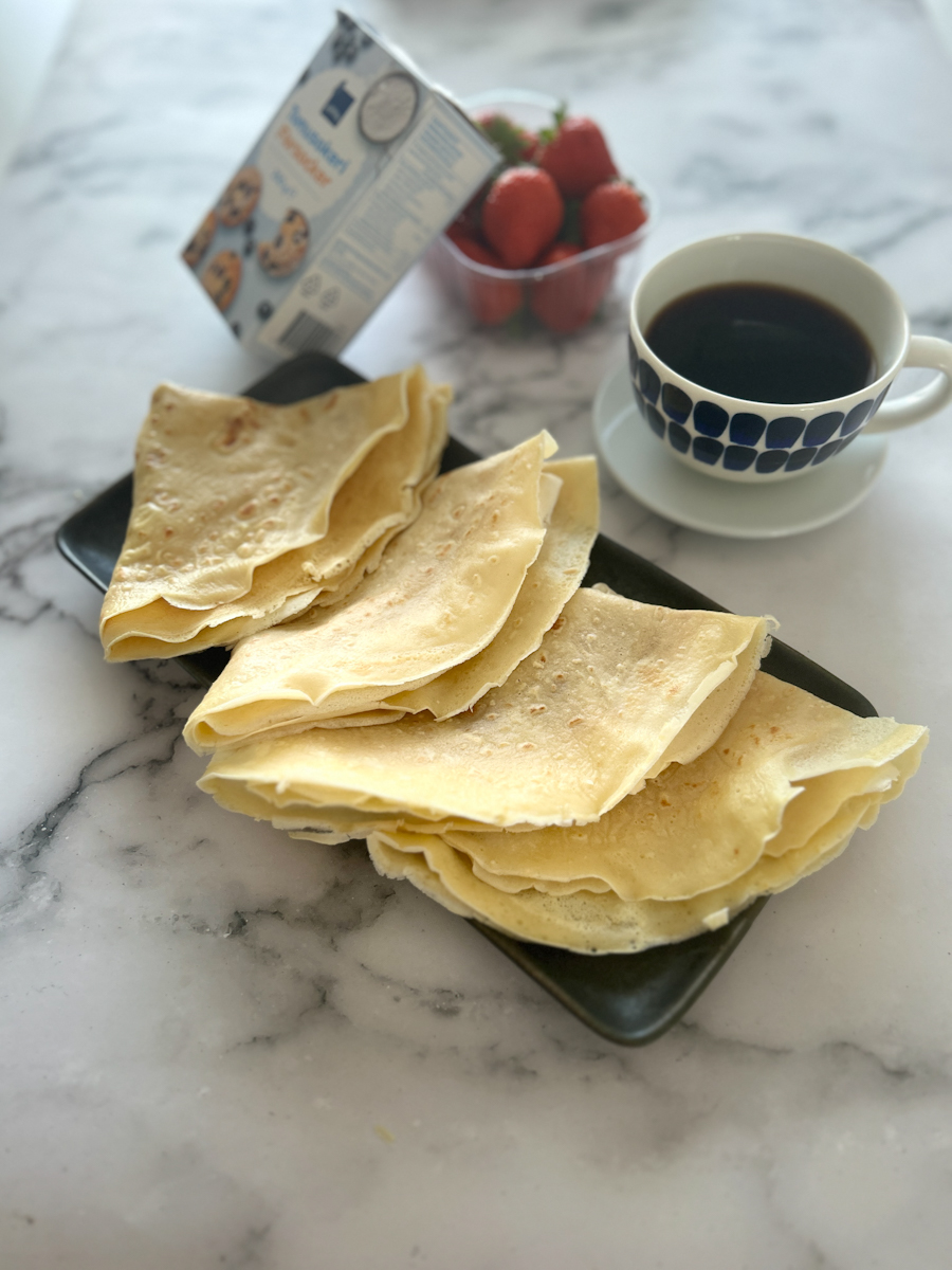 4 crepes folded on a dark blue tray on a table, served with coffee. A powdered sugar package and a box with fresh strawberries are on the table.